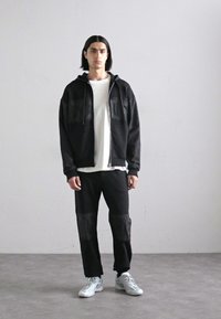 Schwarze Zip-Hoodie mit zwei Brusttaschen, kombiniert mit schwarzen Hosen mit strukturierten Akzenten. Das Modell trägt ein weißes T-Shirt und graue Sneakers.