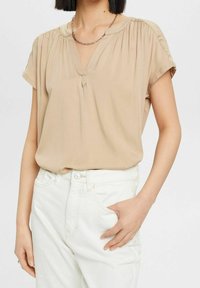 Blouse beige clair à manches courtes, avec un col plissé et une légère découpe en V. Associée à un jean blanc, présentant une coupe décontractée.