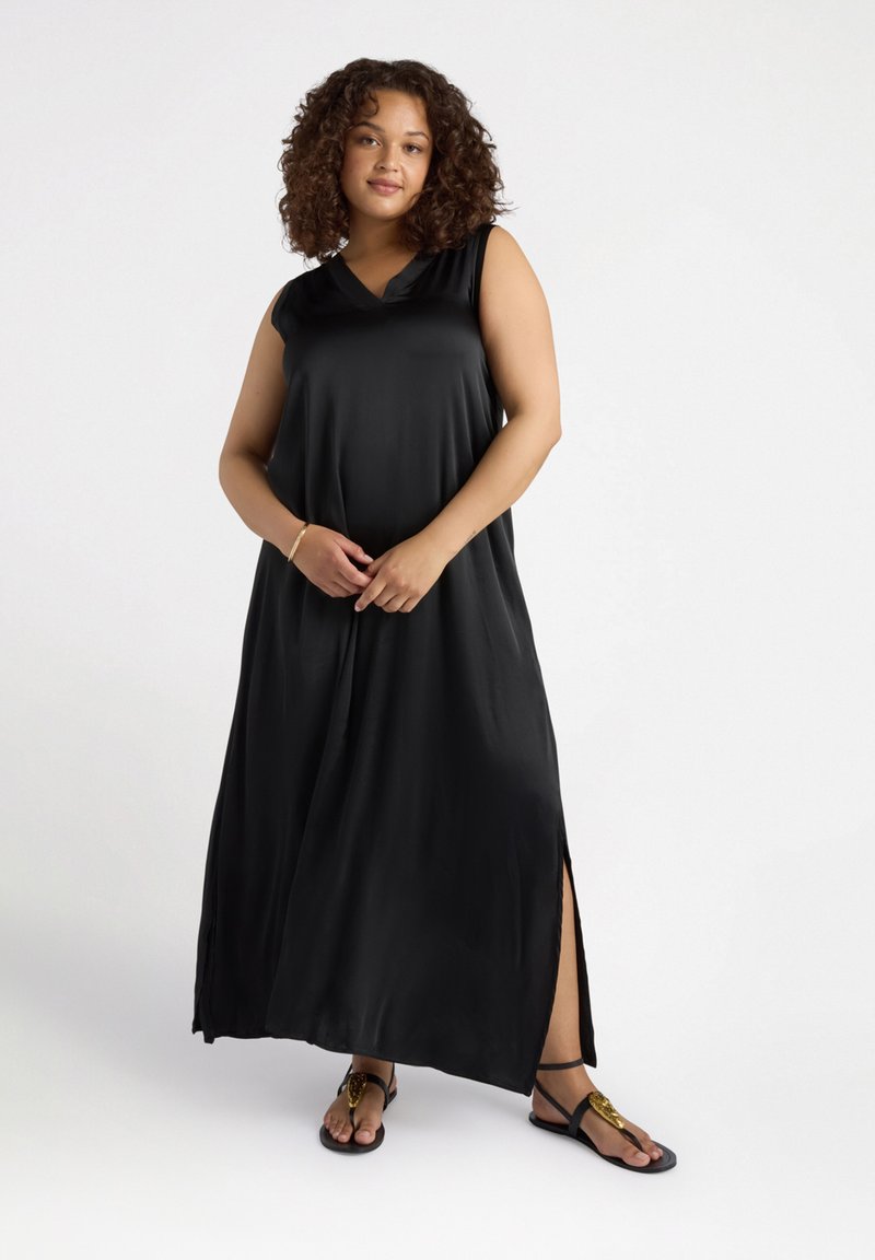 Robe maxi noire sans manches avec un décolleté en V, texture lisse et fentes latérales. Associée à des sandales plates avec des accents dorés.