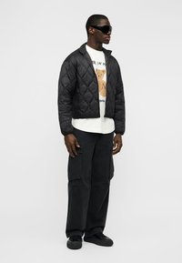 Άνδρας που φοράει μαύρο μπουφάν με καπιτονέ ύφασμα, λευκό t-shirt με στάμπα αρκουδάκι, μαύρο παντελόνι cargo, μαύρα αθλητικά παπούτσια και σκουρόχρωμα γυαλιά ηλίου, στέκεται πλευρικά.