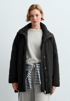 Femme aux cheveux blonds courts porte une veste matelassée noire par-dessus un sweat-shirt gris clair et un vêtement rayé noué autour de la taille.