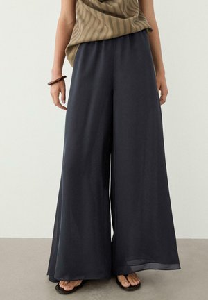 Femme portant un pantalon noir ample à jambes larges, un haut sans manches rayé marron, un bracelet marron et des sandales noires, debout sur un sol clair.