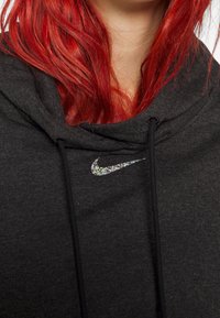Svart hoodie med dragsko, som har en glittrande silvrig Nike-logotyp på bröstet. Tyget ser mjukt ut med en strukturerad yta.