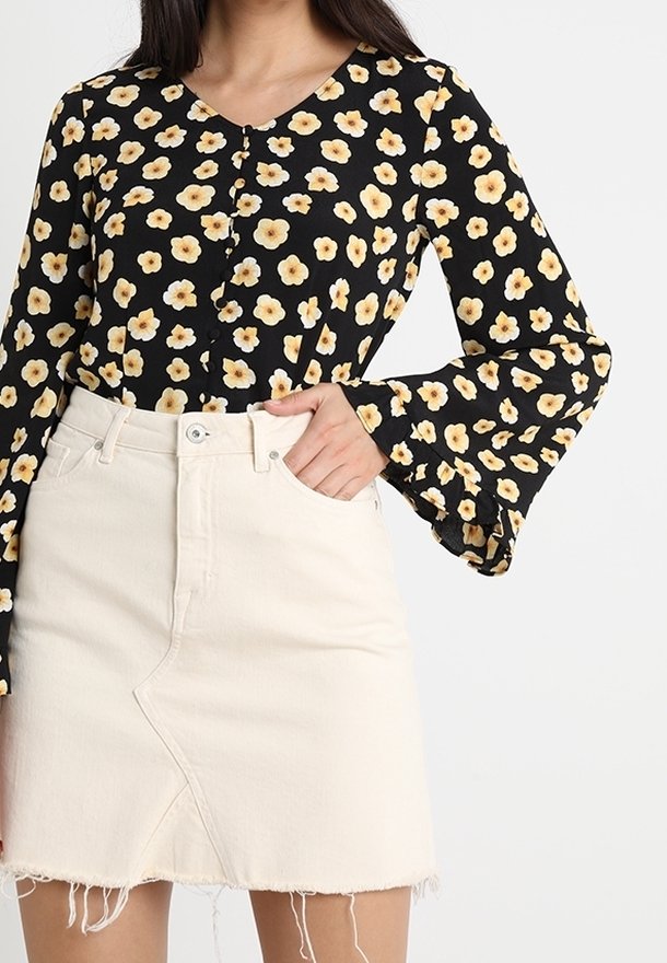Femme portant un haut à manches longues noir avec un motif floral jaune et une jupe en denim beige clair à ourlet effiloché, une main dans la poche.