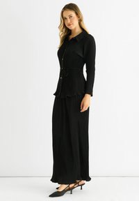 Black pleated dress with a button-down front, long sleeves, tie waist, and scalloped hem. Paired with black pointed-toe slingback shoes.