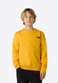 Gul genser laget av mykt stoff med rund hals og ribbestrikkede mansjetter, med en svart logo på venstre bryst.