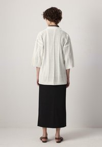 Grosse cardigan en maille blanche à texture côtelée verticale, tissu doux et manches trois quarts, associé à une jupe maxi noire.