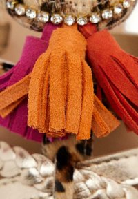 Dettaglio frangia in suede arancione, rosa e rosso con una finitura texturizzata, accompagnato da accenti metallici disposti in un arrangiamento circolare.