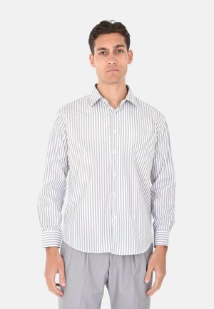 Uomo in piedi, che indossa una camicia a righe verticali bianche e grigie con bottoni e pantaloni grigio chiaro, su uno sfondo chiaro e uniforme.