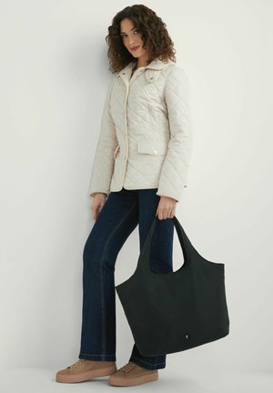 Kobieta w beżowej pikowanej kurtce i ciemnych dżinsach trzyma dużą czarną torbę na ramię, stojąc na białym tle.