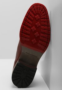 Chaussure en cuir avec une tige marron et une semelle extérieure en caoutchouc noir très texturée. La semelle rouge en caoutchouc présente un motif de bande de roulement en forme de croix.