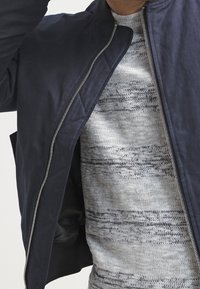 Marineblau bomberjacke mit einem silbernen Reißverschluss, getragen über einem hellgrauen Pullover mit dunkelgrauen horizontalen Streifen und einem strukturierten Muster.