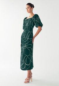 Groene midi-jurk met pofmouwen, voorzien van een wit lijnenpatroon. Gecombineerd met witte sandalen met bandjes en een soepele, aansluitende silhouet.