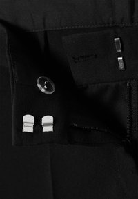 Tissu noir à finition texturée avec un bouton en plastique transparent, une quincaillerie argentée et une poche zippée avec des clips en métal.