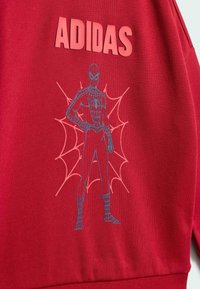 Röd sweatshirt med "ADIDAS" i fet, rosa text. En blå Spider-Man-grafik med ett spindelnätdesign är framträdande.
