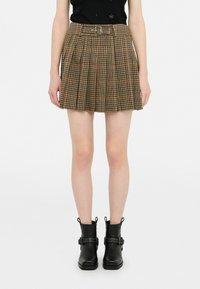 Gonna a pieghe a quadri marrone con cintura, caratterizzata da una vita aderente e un design strutturato. Stivaletti neri alla caviglia con dettagli in metallo completano l'outfit.
