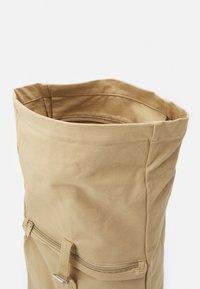 Tan canvas roll-top tas met een ritssluiting, gladde textuur en minimale stikseldetails. Bevat een stevige constructie en functionele vorm.
