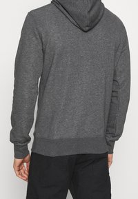 Uomo che indossa un felpa con cappuccio grigio scuro e pantaloni neri, mostrato di spalle su uno sfondo chiaro e semplice.