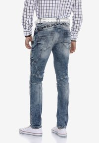 Hellblaue Denimjeans mit schmaler Passform, gestickten Details und mehreren Taschen. Kombiniert mit einem weißen Musterhemd und weißen Sneakers.