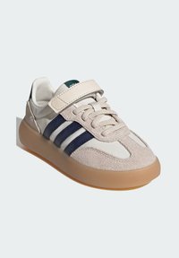 Zapatilla con una parte superior de ante beige y cuero blanco, con franjas marinas, una correa de velcro y una suela de goma translúcida.