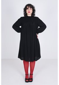 Zwarte smockmaxi-jurk met lange mouwen en een gerimpelde halslijn, gecombineerd met rode panty's en zwarte platform sandalen.