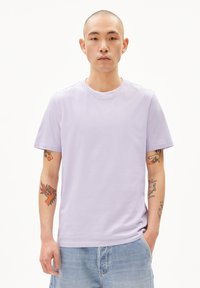 Lavendelfarbenes Rundhals-T-Shirt aus weichem Baumwollmaterial, mit kurzen Ärmeln und einem lässigen Schnitt. Getragen mit hellen Jeansshorts und sichtbaren Tattoos.