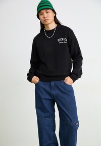 Black knit sweater with logo on the chest, paired with loose blue denim pants featuring patchwork details and a logo patch on the leg.