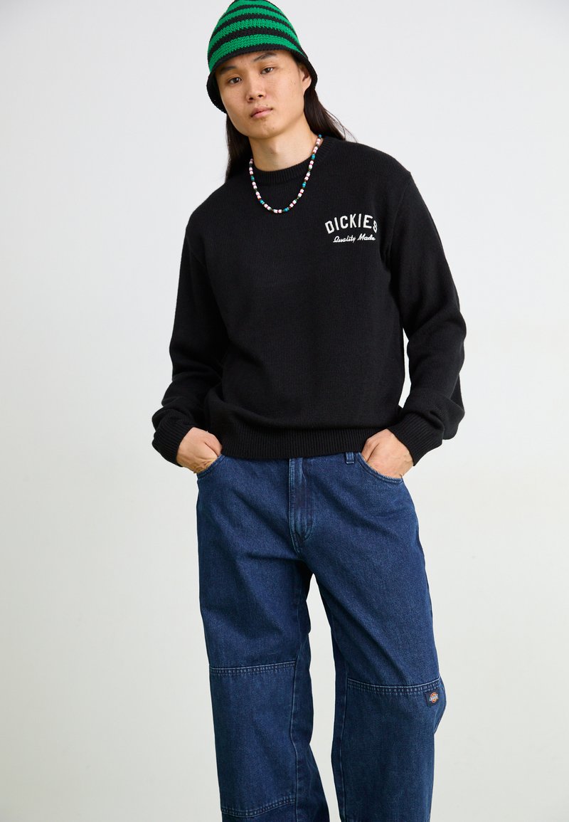 Pull en maille noir avec logo sur la poitrine, associé à un pantalon en denim bleu clair large avec des détails en patchwork et un patch logo sur la jambe.