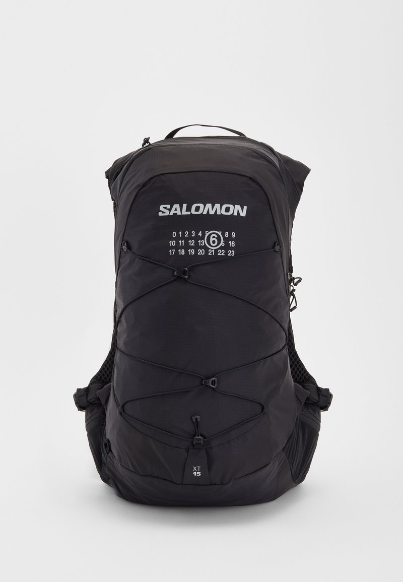 Mochila preta com tecido texturizado, alças ajustáveis e um logótipo proeminente. Apresenta cordas elásticas na frente e dois bolsos laterais.