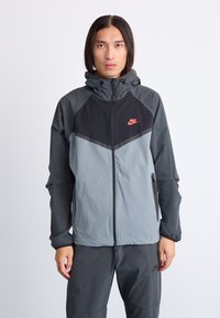 Nike windbreaker jacket in two-tone gray and black. Features a zip front, drawstring hood, and side zip pockets. Soft, lightweight material.