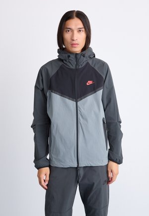 Veste coupe-vent Nike en gris et noir bicolore. Dotée d'une fermeture éclair à l'avant, d'une capuche avec cordon de serrage et de poches latérales zippées. Matériau doux et léger.