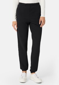 Sorte sweatpants med elastisk talje, tætsiddende pasform og manchetter, parret med hvide sneakers. Blød stoftekstur synlig.