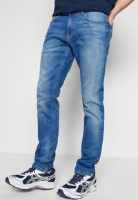 Jeans de mezclilla azul con corte recto, que cuentan con cinco bolsillos y una cremallera clásica. Combinados con zapatillas de correr blancas y marinas.
