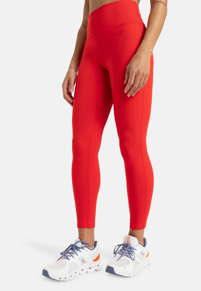 Leggings rouges taille haute en matériau extensible, présentant une texture lisse et un design ajusté, associés à des chaussures de sport blanches.