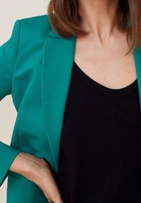 Turkusowa marynarka w dopasowanym kroju, z wyrazistymi klapami i gładką fakturą, noszona na czarnym topie na ramiączkach. Ręka spoczywa na biodrze.