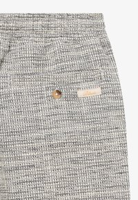 Un pantalon en tissu texturé présentant un mélange de fils crème et marine, avec une fermeture à bouton et une petite étiquette de marque beige.