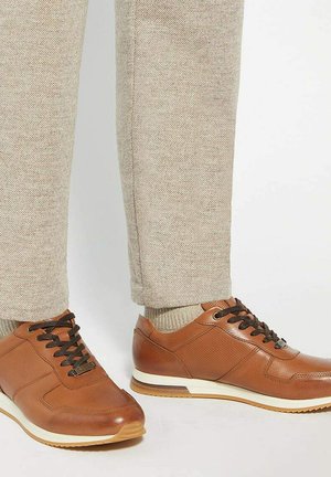 Brown leather sneakers with black laces worn with beige pants and matching beige socks, standing on a white floor.