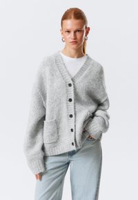 Cardigan en tricot gris clair avec col en V, boutons sur le devant, bords côtelés et une seule poche plaquée. Texture douce, coupe oversize.