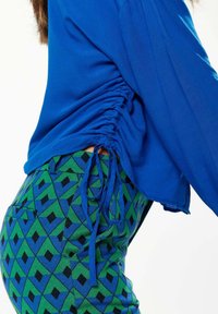 Blue long-sleeve top with gathered side detailing paired with green and blue patterned pants featuring geometric shapes in black accents.