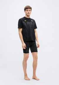 Μαύρο αθλητικό σύνολο που αποτελείται από κοντομάνικη μπλούζα με υφή και σορτς, και τα δύο με διακριτικά μοτίβα. Η μπλούζα έχει χρυσές λεπτομέρειες.