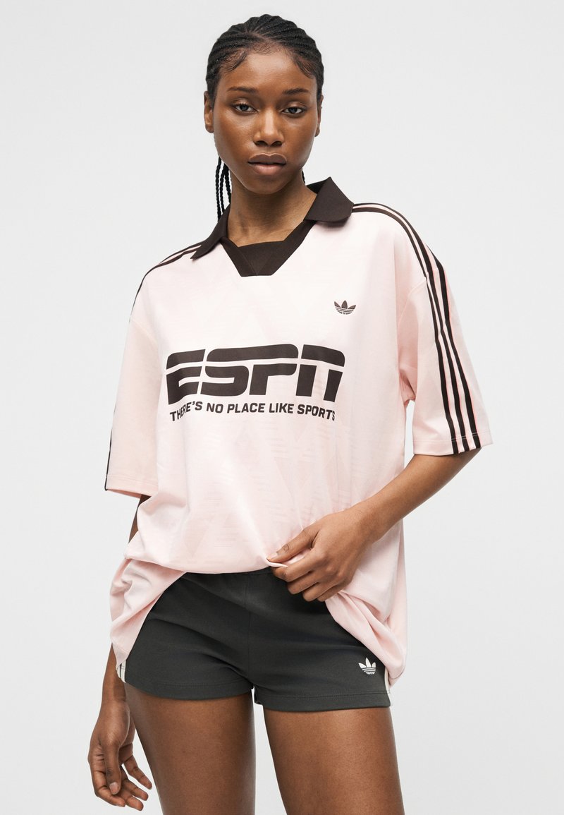 Femme portant un maillot de sport ample rose clair avec un col noir, le texte ESPN noir, et un short foncé avec le logo Adidas sur la jambe gauche.