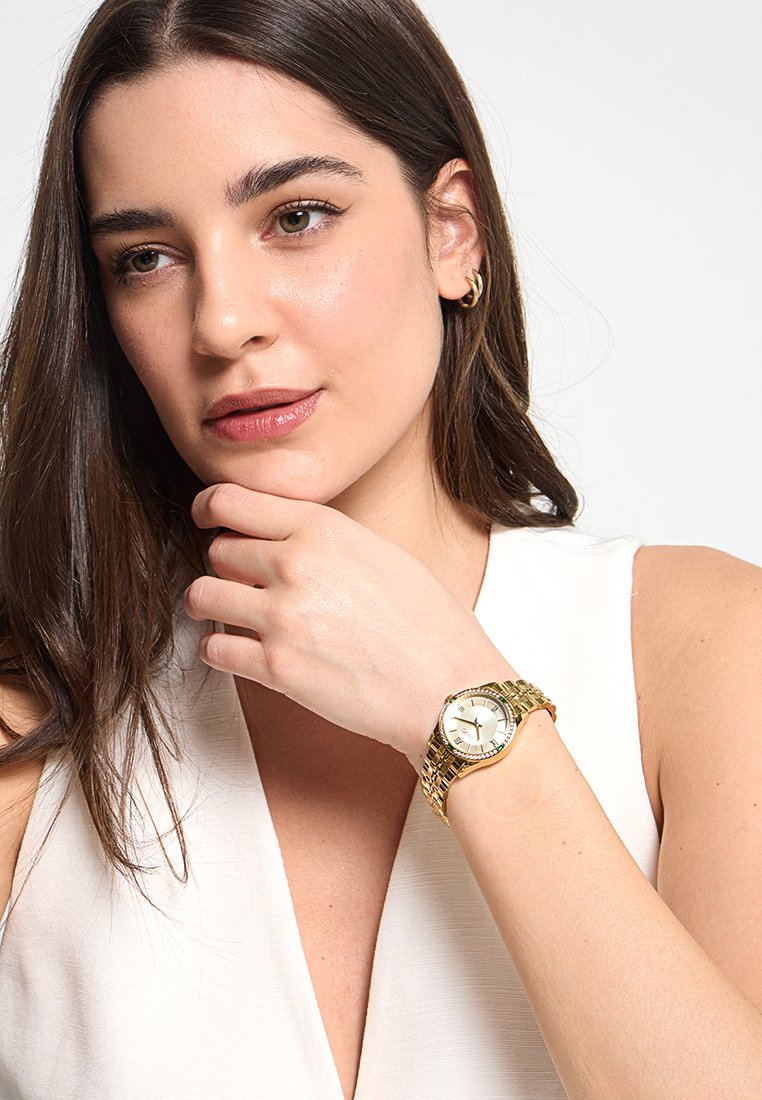 Goudkleurig polshorloge met een ronde wijzerplaat, witte dial en een gouden band met een gestructureerd oppervlak. De hand van het model rust op haar kin.