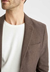 Uomo con un blazer marrone testurizzato sopra una maglietta bianca a collo rotondo, mostrato dalla collana fino a metà torso su uno sfondo neutro.