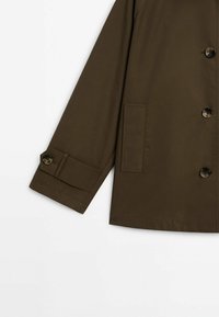 Tumma oliivinvihreä takki, joka on valmistettu sileästä kankaasta; siinä on kuusi nappia, rintatasku ja napilla suljetut hihansuut. Yksinkertainen, jämäkkä muotoilu.