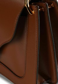 Bruine leren handtas met een gladde textuur, voorzien van een uniek gebogen ontwerp, gouden beslag en contrasterende stiksels langs de randen.