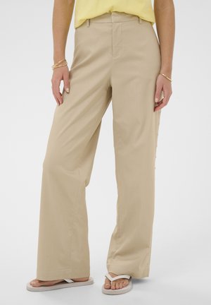 Naine, kes kannab beežikaid laiade säärega pükse, kollast pluusi, kuldseid käevõrusid ja valgeid plätusid, seisab ühtlasel heledal taustal.