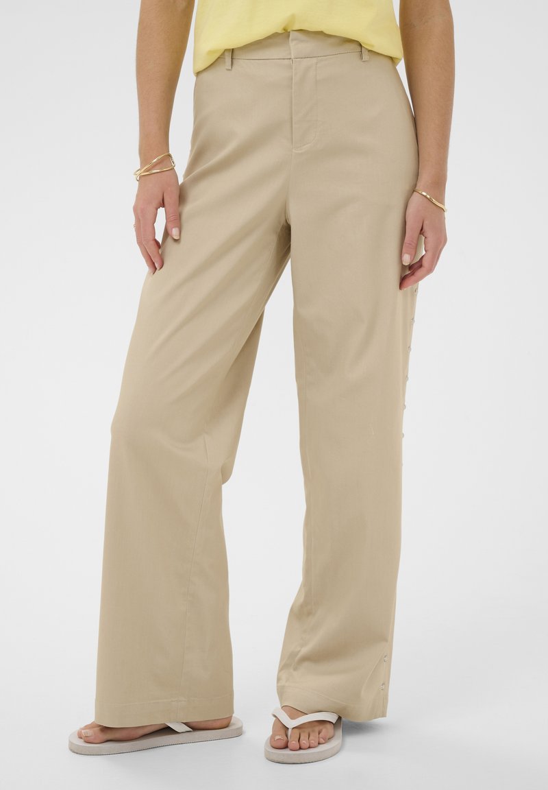 Femme portant un pantalon large beige, un haut jaune, des bracelets dorés et des tongs blanches, debout sur un fond clair uni.