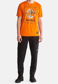 Hombre vistiendo una camiseta brillante naranja de Timberland con el logo de un árbol, pantalones cargo negros y botas negras de cordones, de pie contra un fondo blanco.
