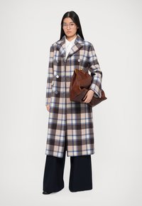 Donna che indossa un lungo cappotto a quadri nei colori beige, blu e marrone, mentre tiene una grande borsa di pelle marrone, con pantaloni larghi neri e occhiali.