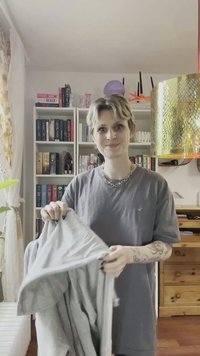 Svijetlosivi sako drži osoba u sivom majici. Sako ima jednostavan dizajn i zatvaranje na patentni zatvarač. U pozadini su police s knjigama.